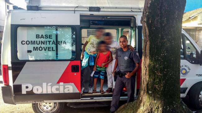 Imagem mostra Ronie sorrindo, vestido de farda de policial. Ao lado dele, três crianças posam para foto em cima de van da polícia, uma base comunitária móvel