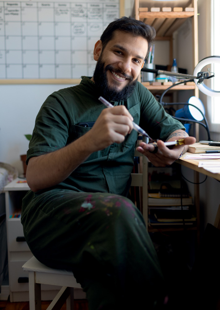 Homem de barba grande sorri em mesa de ateliê, segurando caneta e papel. Veste camisa escura.