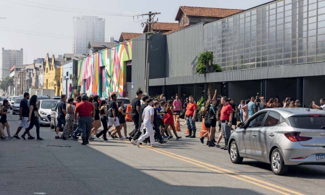 Imagem mostra várias pessoas atravessando avenida, com carros parados esperando os pedestres. Ao fundo, galpão preto com grafites coloridos