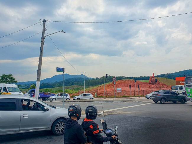Imagem de rua mostra dois motoqueiros em primeiro plano, olhando para a frente, e outros carros parados em semáforo. Ao fundo, terreno com terra revirada e um trator e, mais ao fundo ainda, o Pico do Járagua