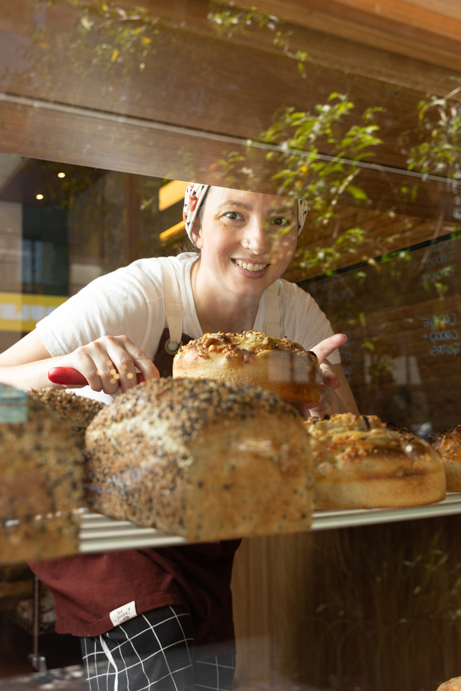 Atrás de um vidro, uma mulher sorri para a foto. Tem uma prateleira com pães na sua frente.