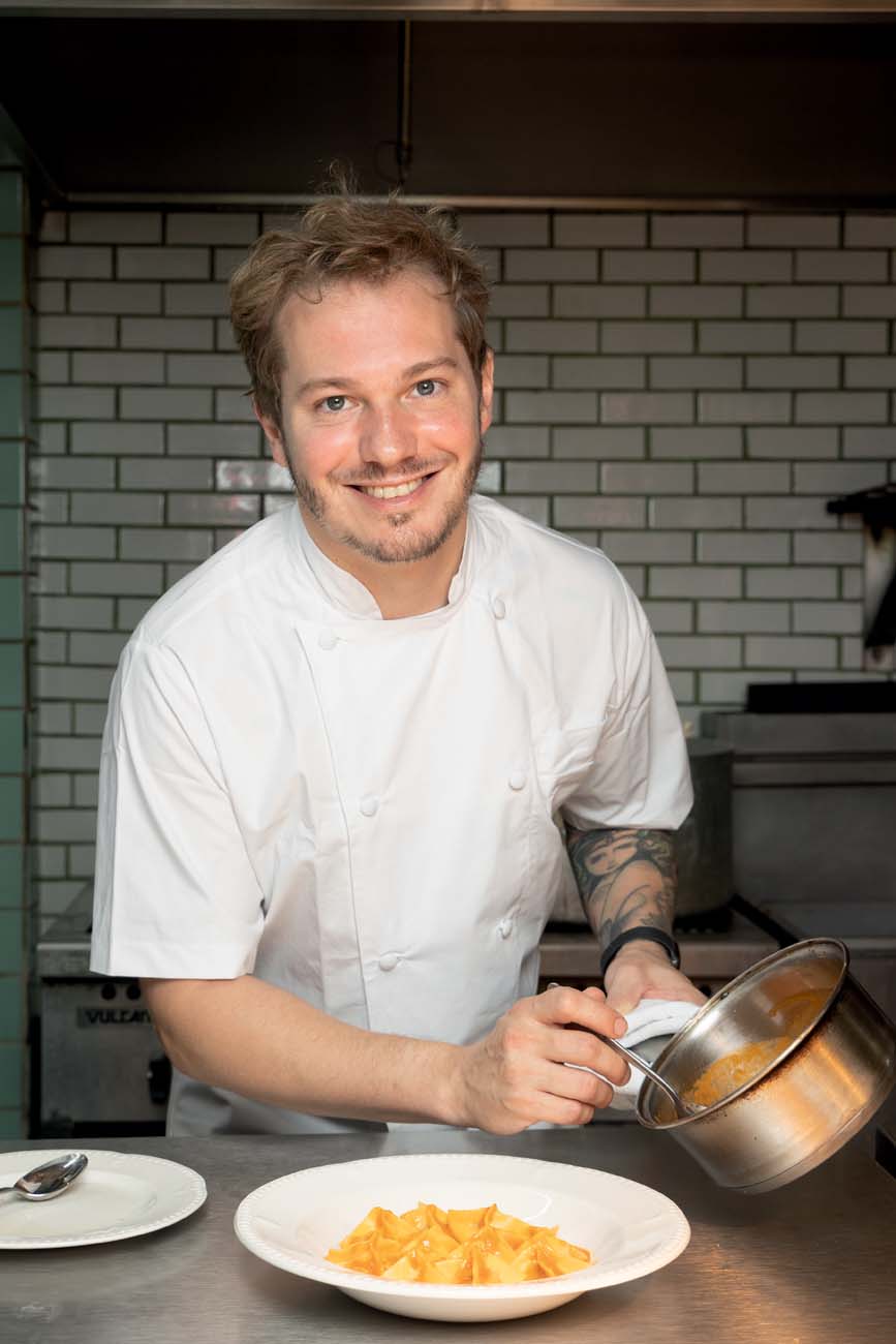 chef loiro de dólmã sorrindo para a foto segurando panela de cobre com massa em prato fundo de louça branca na frente.