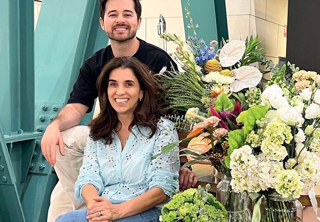 Homem e mulher posam lado a lado, ele atrás dela, que está sentada ao lado de grande arranjo floral. Ela veste jeans e camisa azul, ele camiseta preta.