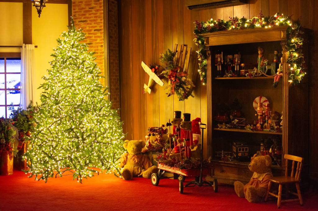 Parte de uma sala de estar com piso vermelho e uma grande árvore de natal com luzes à esquerda. À direita tem uma estante de madeira com várias decorações de natal.