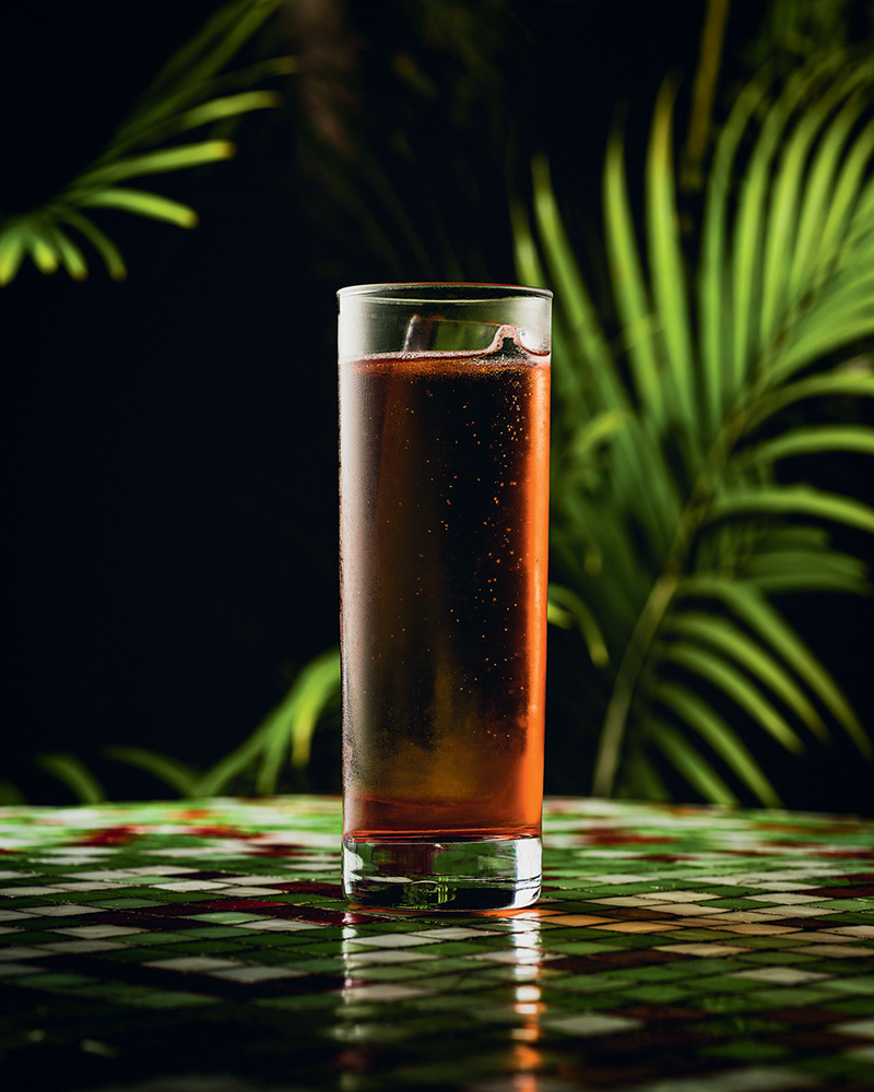 Drinque avermelhado no copo longo com plantas atrás em fundo preto