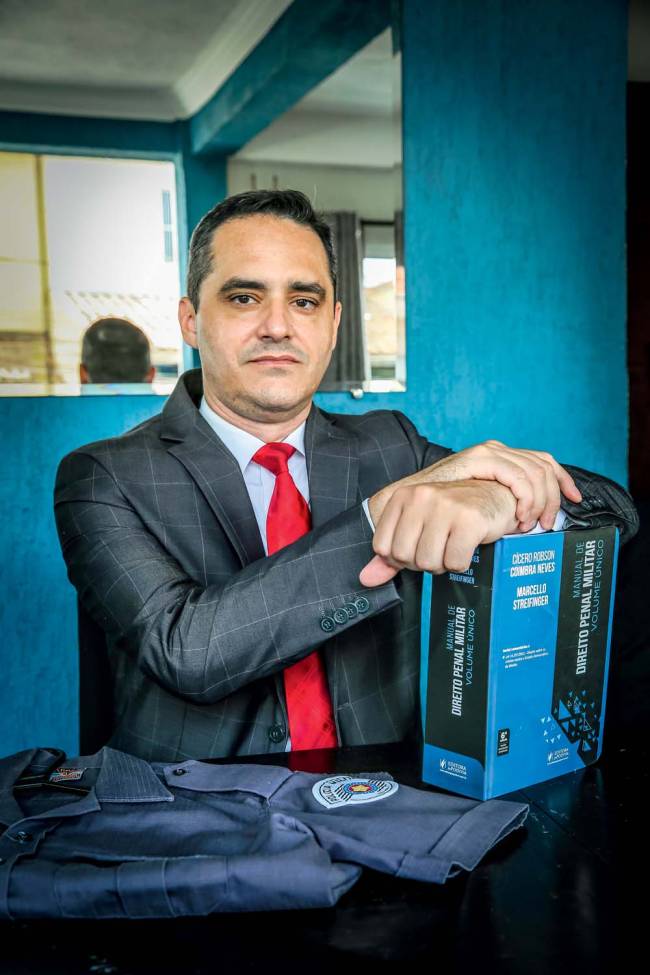 Imagem mostra Thiago sentado de terno e gravata, com as mãos em cima de um grosso livro de direito militar e, ao lado, uma farda da PM