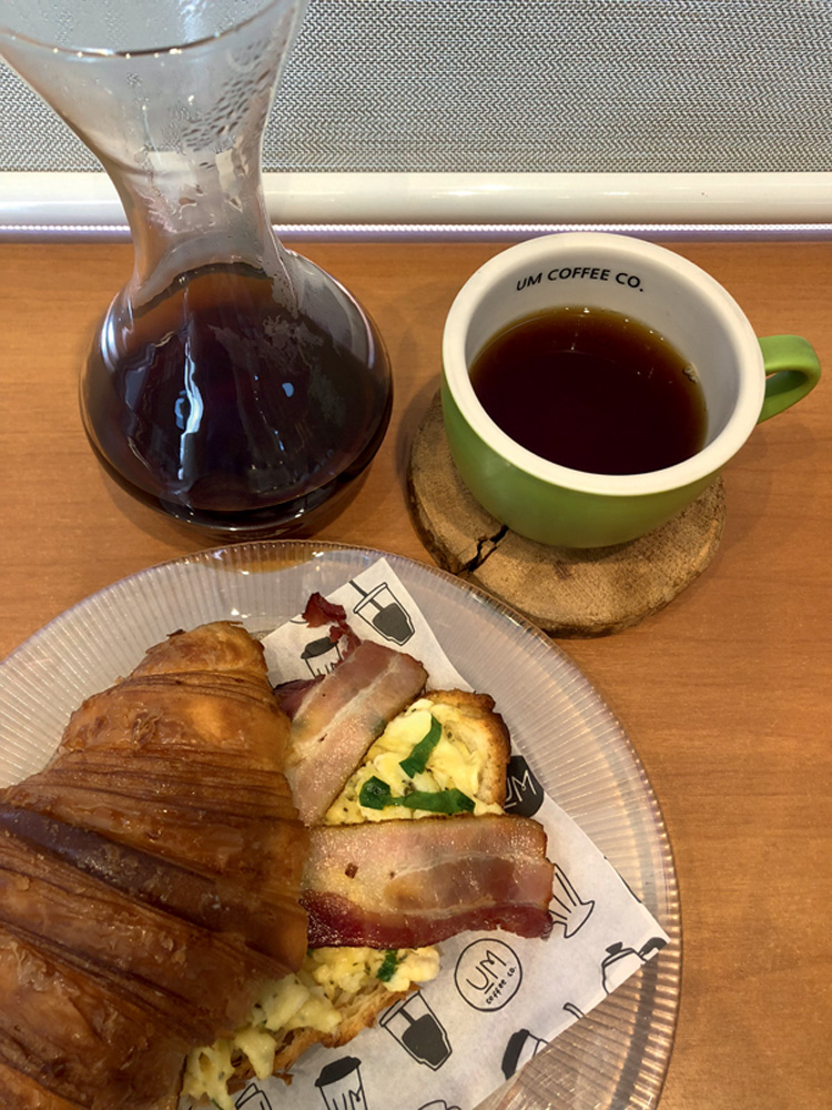 Foto aérea de croissant com ovos mexidos e fatias de bacon e xícara de café coado ao lado de despenser com café