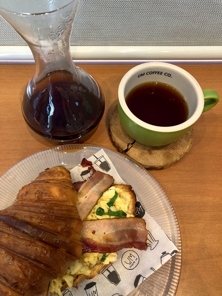 Circuito Comer & Beber – Um Coffee Co. Foto aérea de croissant com ovos mexidos e fatias de bacon e xícara de café coado ao lado de despenser com café