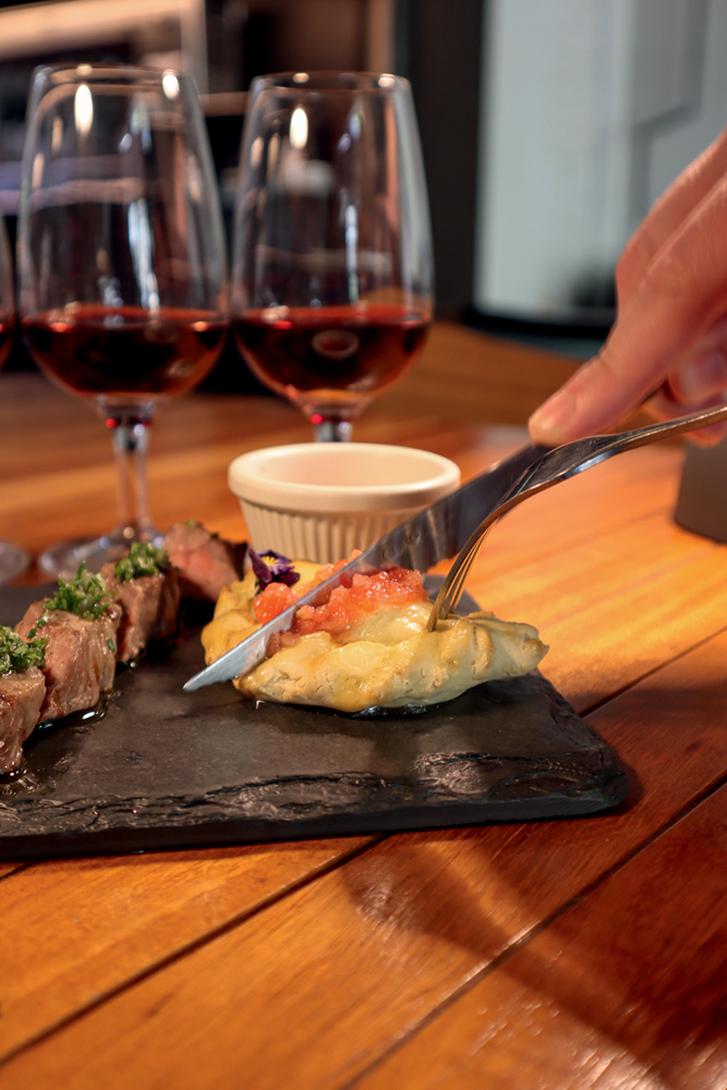 Circuito Comer & Beber – Bardega Talhares cortando empanada com duas taças de vinho tinto ao fundo