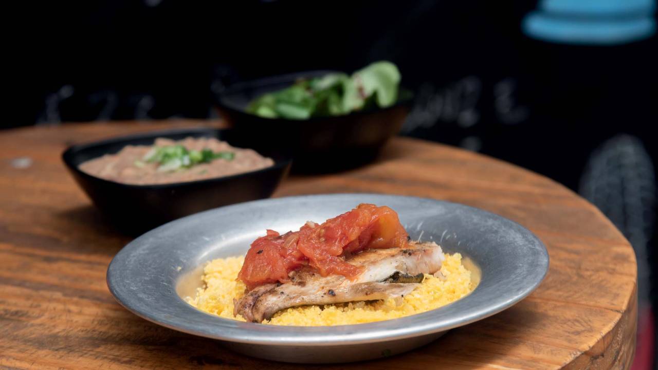 Sobre uma mesa de madeira, um prato de alumínio com cuscuz de milho embaixo, pedaço de peixe por cima coberto de molho de tomate.