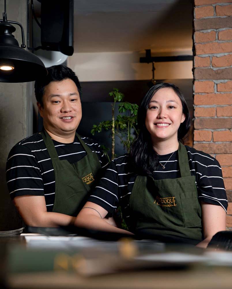 Casal de chefs asiáticos com braços apoiados em bancada usando camisa preta com listras brancas e avental verde escuro.