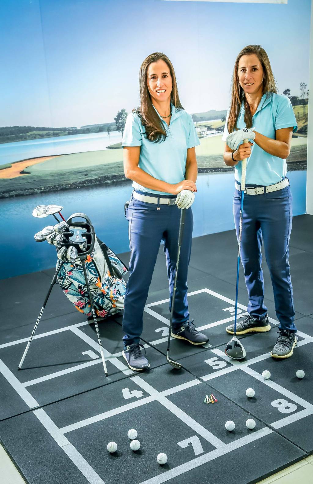 Gêmeas de cabelos castanhos lisos posam de pé segurando taco de golfe em ambiente com fundo de tela que imita um campo.