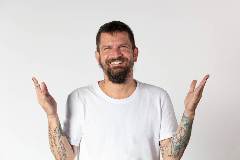 Homem branco de barba posa com os braços abertos, sorrindo e vestindo camiseta branca.