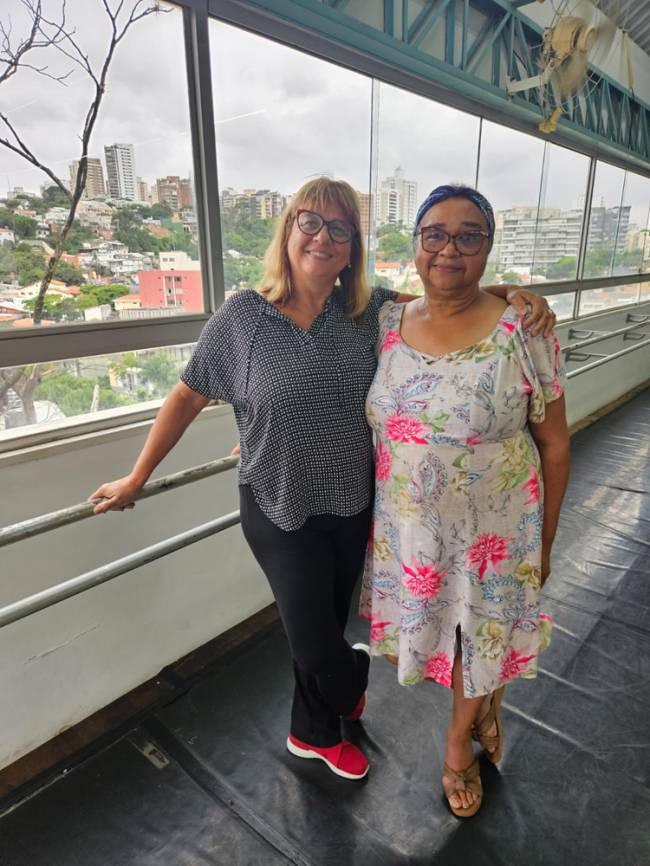 Imagem mostra duas mulheres sorrindo abraçadas com janela aos fundos. À direita, uma é loira e usa camiseta e calça e a da esquerda usa um vestido florido