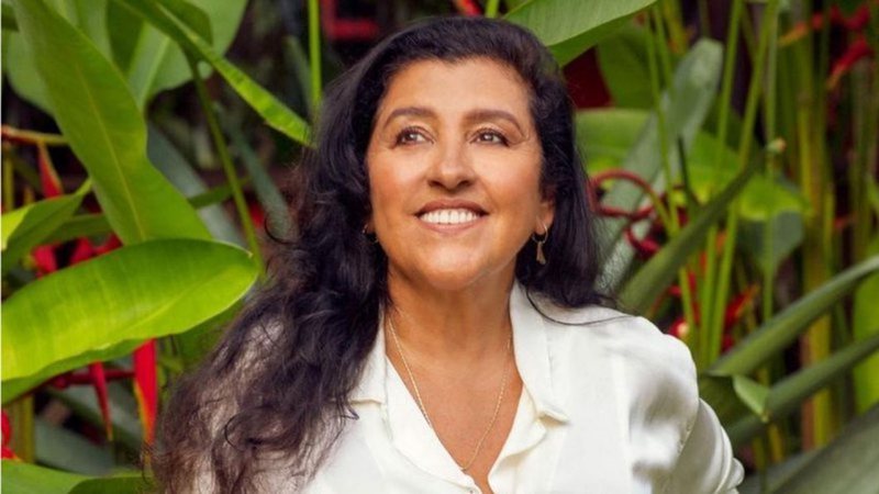 Imagem mostra Regina Casé sorrindo. Ela usa camisa branca e está em local com fundo cheio de plantas verdes