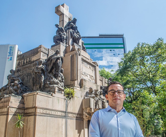 Homem branco grisalho e cacreca de roupas pretas posa em frente a mesa com livro grande. Atrás, prateleiras exibem mais livros