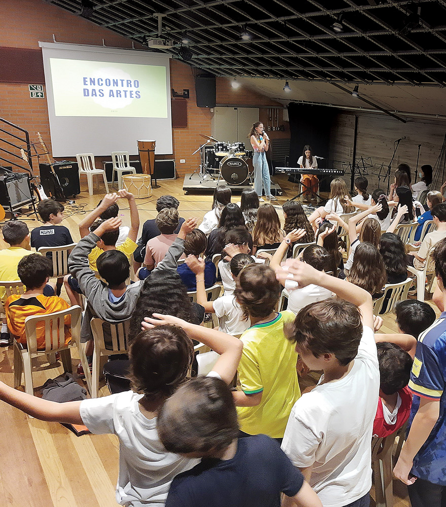 Imagem mostra vários alunos olhando palco em que aparece uma menina cantando e outra tocando piano. Ao fundo, pode-se ler no telão 