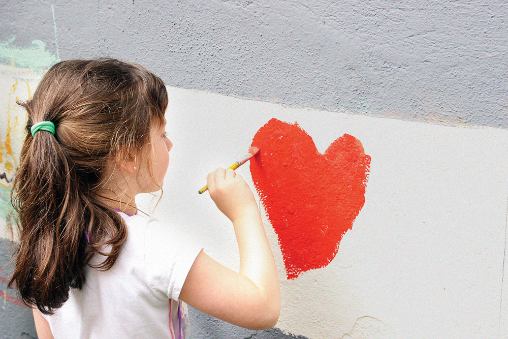 Imagem mostra aluna pintando coração vermelho com tinta em parede