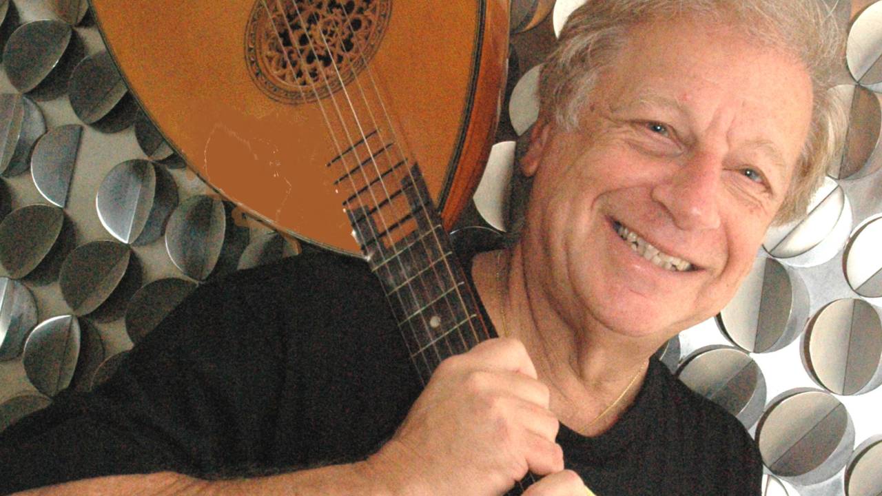 Homem grisalho sorri com instrumento de cordas apoiado no ombro