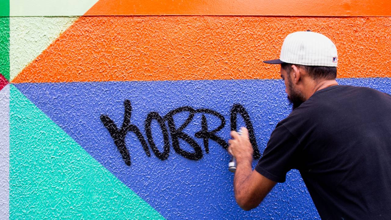 Artista grafiteiro posa de costas assinando "Kobra" em parede nas cores azul, verde e laranja. Ele usa camiseta preta e boné branco