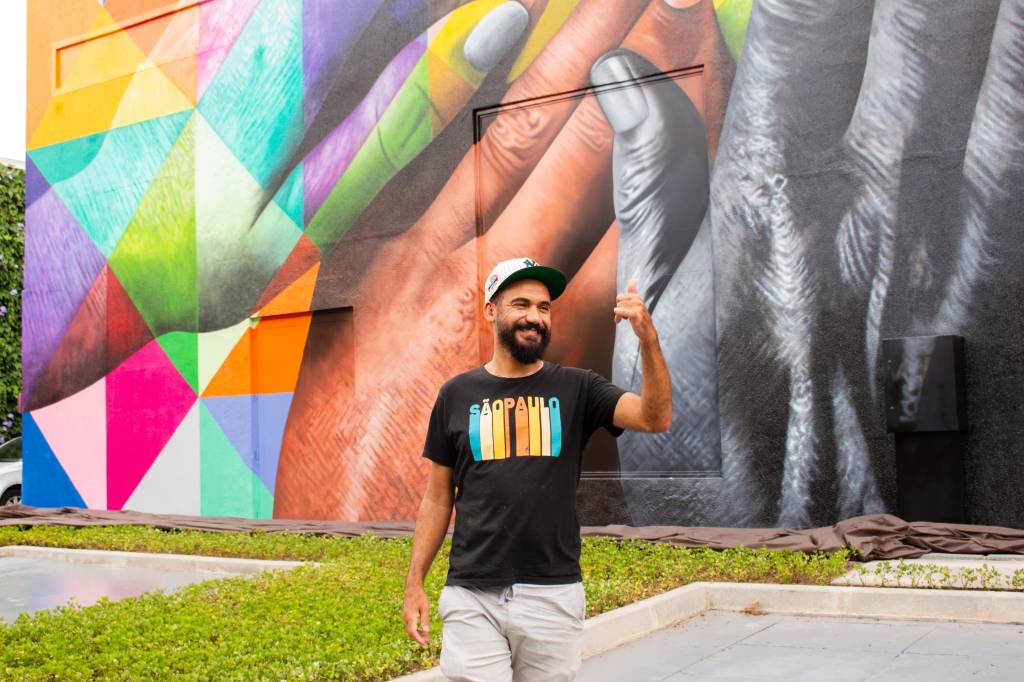 Artista com barba, camiseta preta com estampa colorida e shorts brancos posa com uma mão para cima. Ele está em frente a grande mural em área externa, com arte colorida que exibe mãos unidas em vários tons