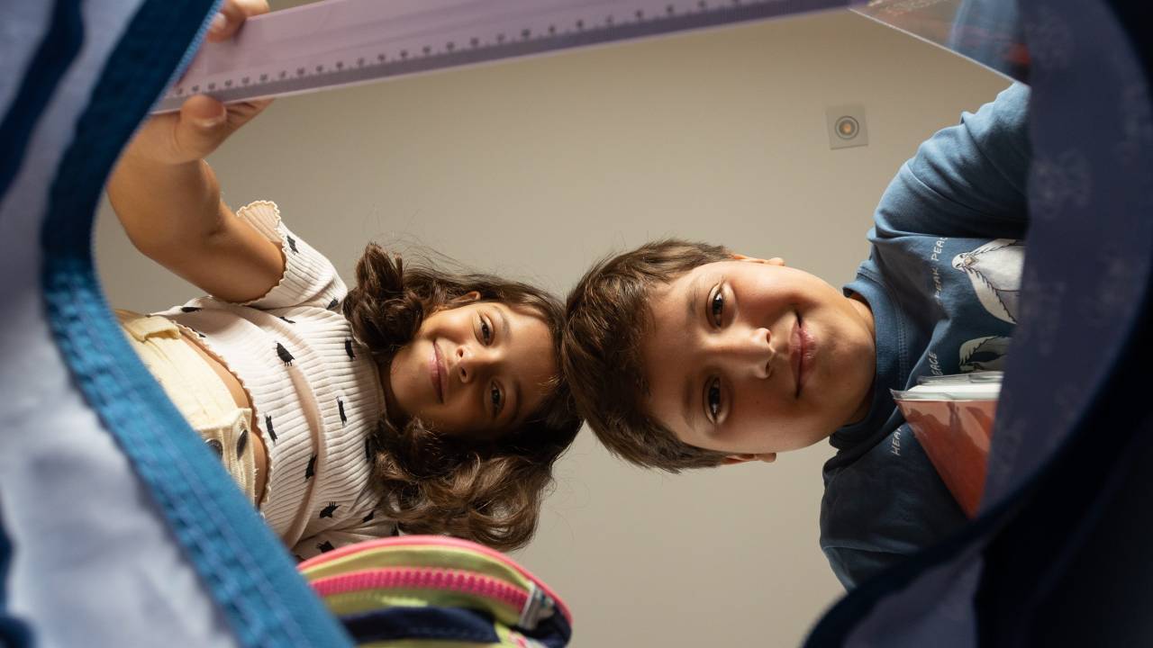 Imagem mostra crianças olhando para câmera, que está dentro de mochila escolar. Imagem mostra abertura de mochila e, ao fundo, as duas crianças sorrindo, segurando réguas e estojo