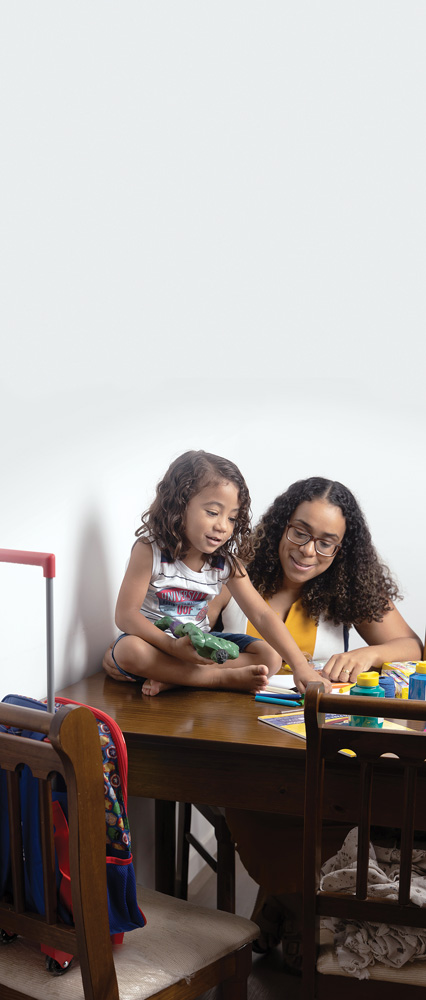 Imagem mostra criança em cima de mesa segurando boneco e apontando para lápis de cor e tintas. Junto, mãe, sentada em cadeira na mesa, que segura o filho pelas costas