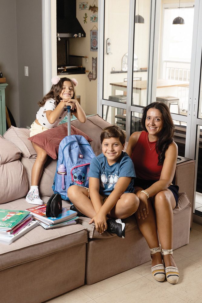 Imagem mostra mãe e duas crianças (uma menina e um menino) em sofá de apartamento, com livros escolares e mochila escolar ao lado. Ao fundo, sacada, com churrasqueira