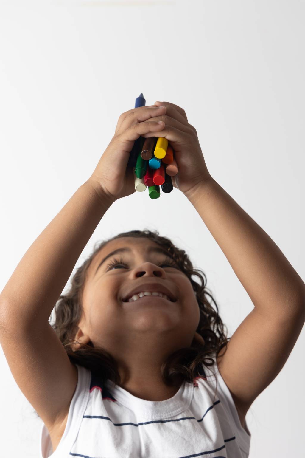 Imagem mostra criança sorrindo, segurando lápis de cor entre as mãos e os erguendo para cima