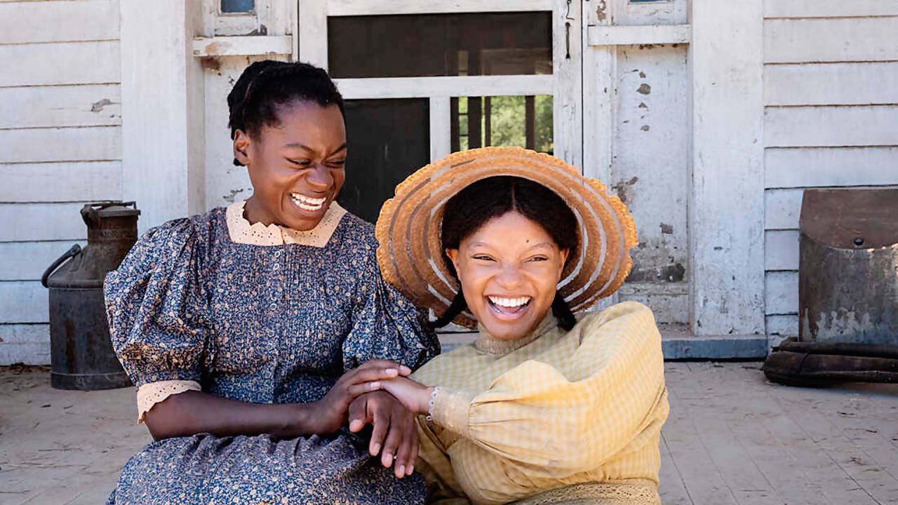 Foto exibe duas mulheres negras com vestidos antigos em escada de terraço de casa com madeira branca. A da direita usa chapéu de palha