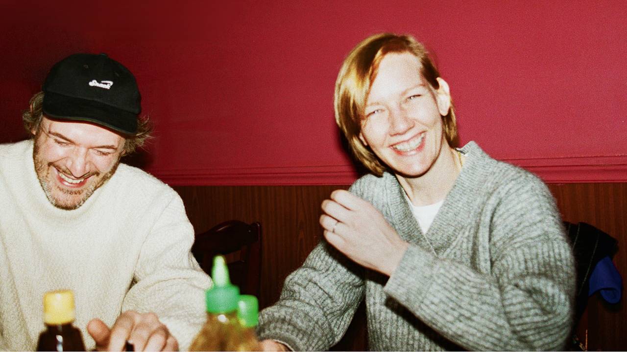 Um homem e uma mulher, ambos brancos, dão risada em mesa de restaurante. Ele, à esquerda, veste boné e suéter branco e ela uma blusa cinza
