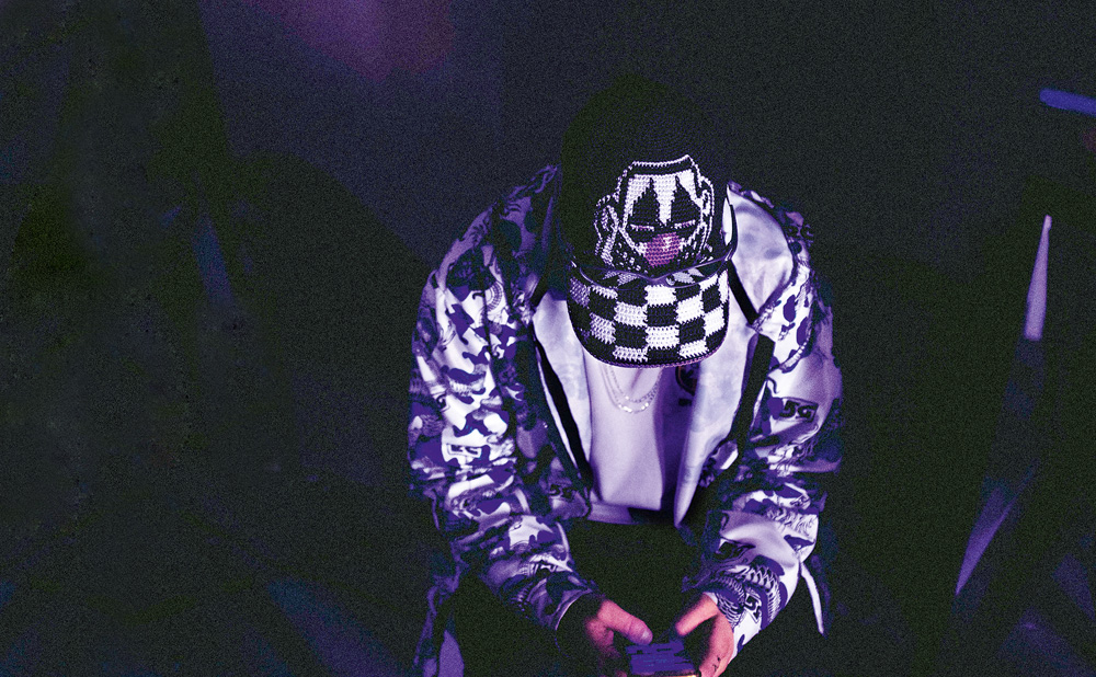 Imagem mostra DJ com a cabeça abaixada, mostrando boné com desenho de palhaço e aba quadriculada em ambiente escuro e arroxeado