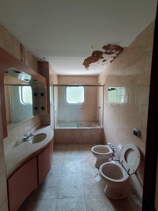 Imagem mostra banheiro com chão e pia de pedra clara. Ao fundo, boxe com banheira e ao lado, pia com espelho. Acima, teto, com a parte direita, em cima do vaso sanitário e do bidê, com manchas escuras