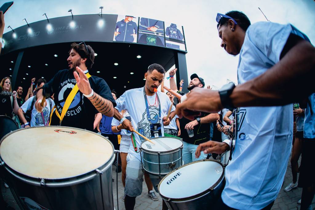 Imagem mostra bateria tocando em espaço aberto, com três pessoas em primeiro plano, uma com um grande surdo, outra com uma ripa e outra com uma caixa. Todos são homens