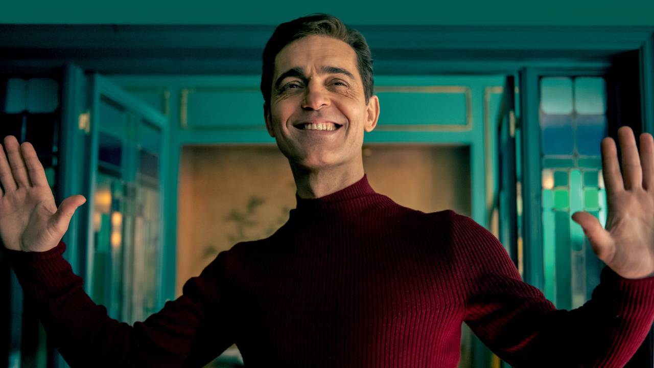Homem branco sorri de braços abertos em frente a parede e porta verdes, vestindo suéter vermelho escuro