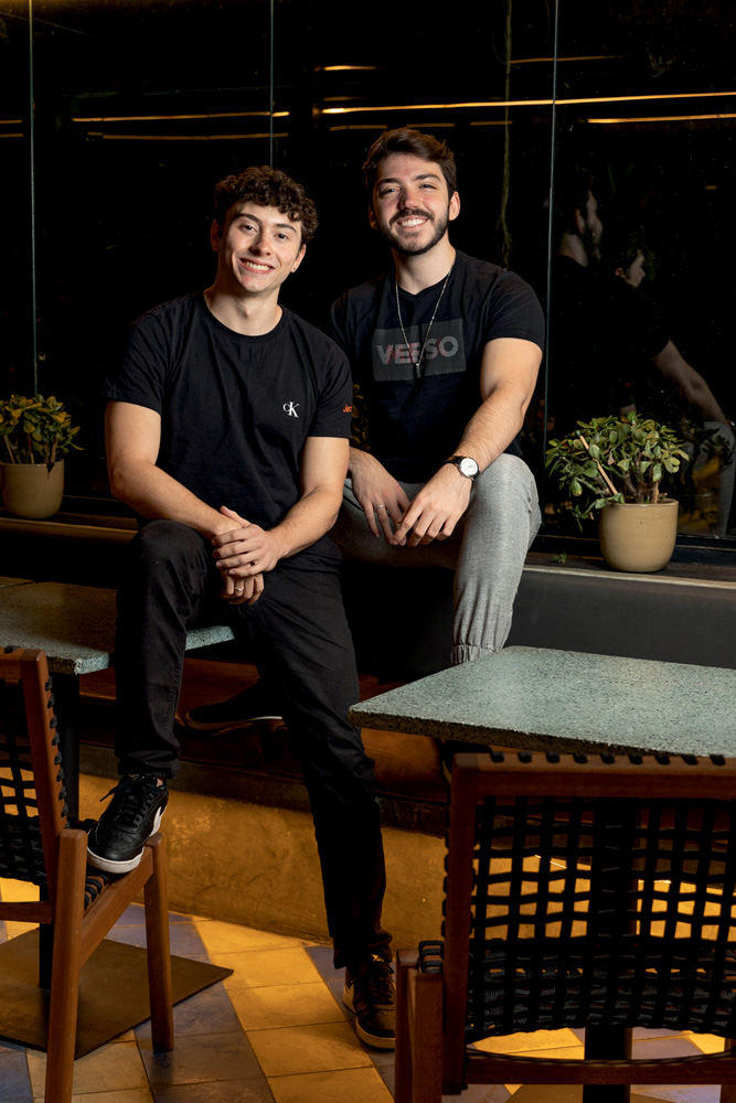 Imagem mostra Guilherme e Lucas sentados em mesa de restaurante, sorrindo. Eles usam camisa preta e calças jeans