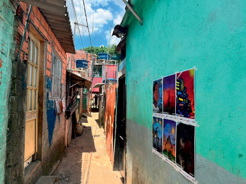 Imagem mostra seis cartazes com artes colados em muro de viela com casas de madeira e alvenaria em rua com chão parcialmente de terra