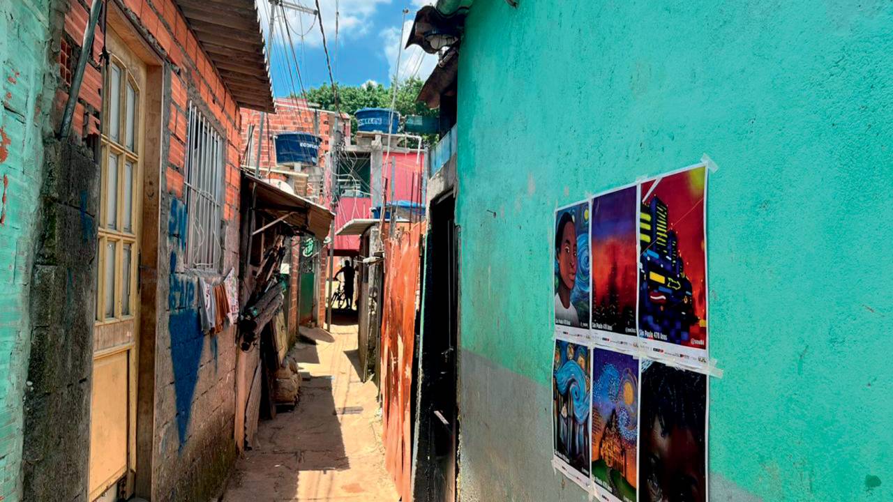 Imagem mostra seis cartazes com artes colados em muro de viela com casas de madeira e alvenaria em rua com chão parcialmente de terra
