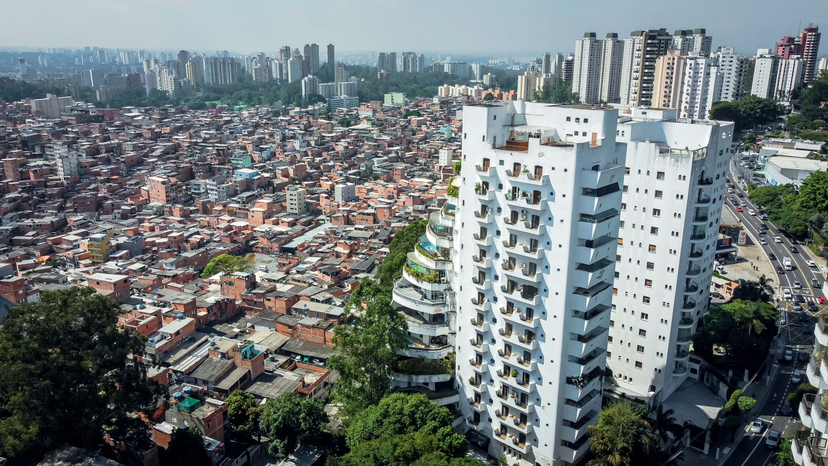 Imagem mostra foto aérea de prédio com uma piscina por andar, com sacadas em espiral e, ao lado, favela, com pequenas casas, a maior parte com tijolos expostos. No canto direito, grande avenida movimentada