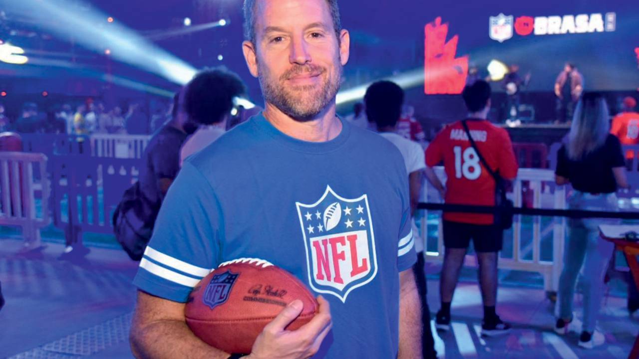 Homem branca de barba posa com bola de futebol americano no colo. Veste camiseta da NFL azul.