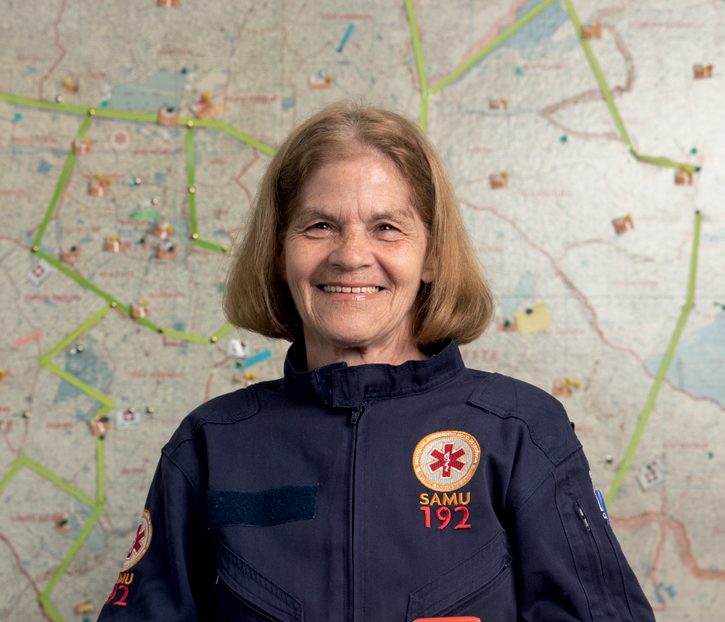 Imagem mostra mulher sorridente com uniforme que pode-se ver o logo do Samu e os números de telefone do serviço, 192. Ela segura uma pequena ambulância, também com o nome e telefone do serviço. Ao fundo, um mapa da cidade de São Paulo, que ocupa todo a parede