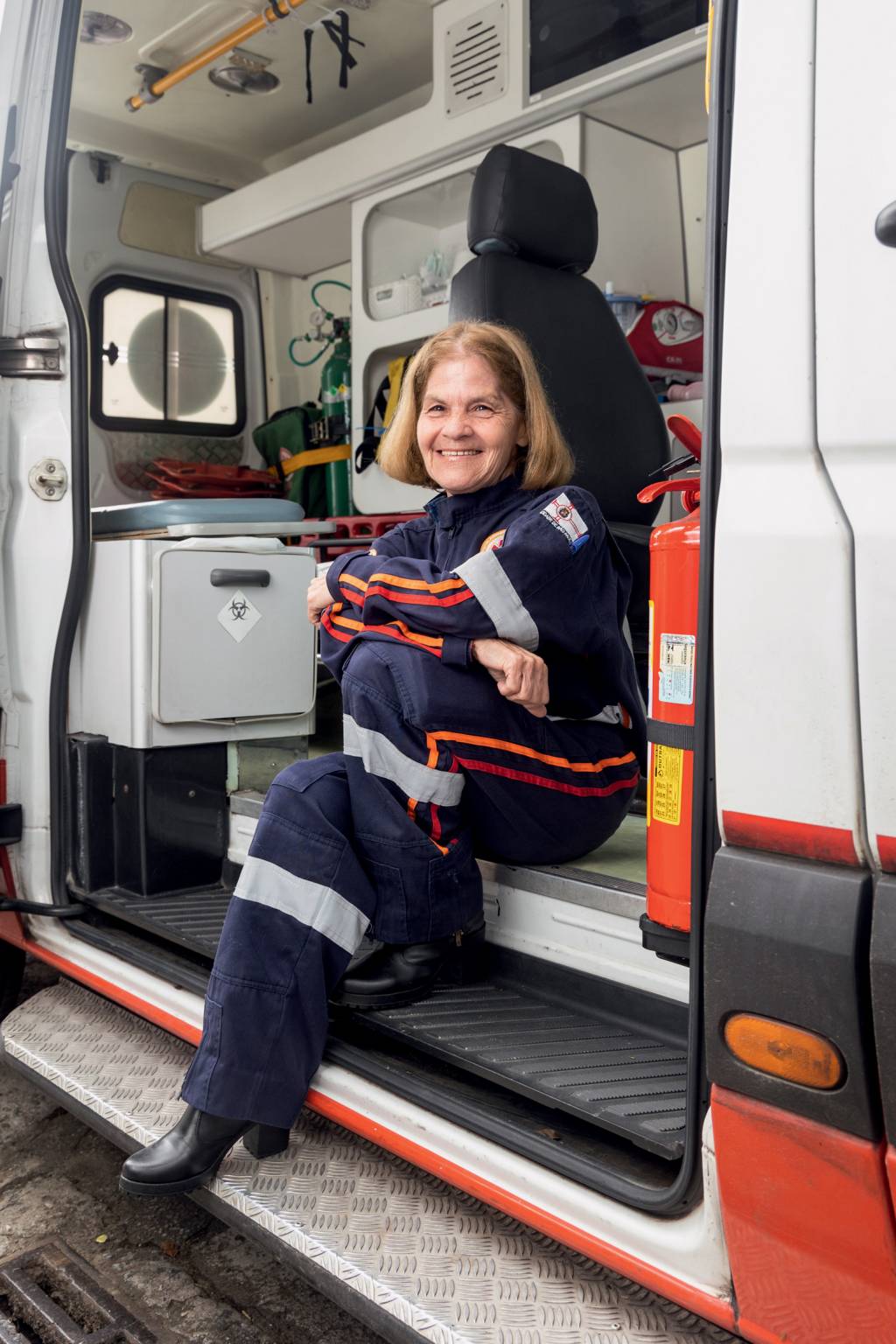 Imagem mostra Maria Jalva sentada em ambulância, que tem porta lateral aberta. Ela sorri de dentro da ambulância, que é cheia de equipamentos médicos