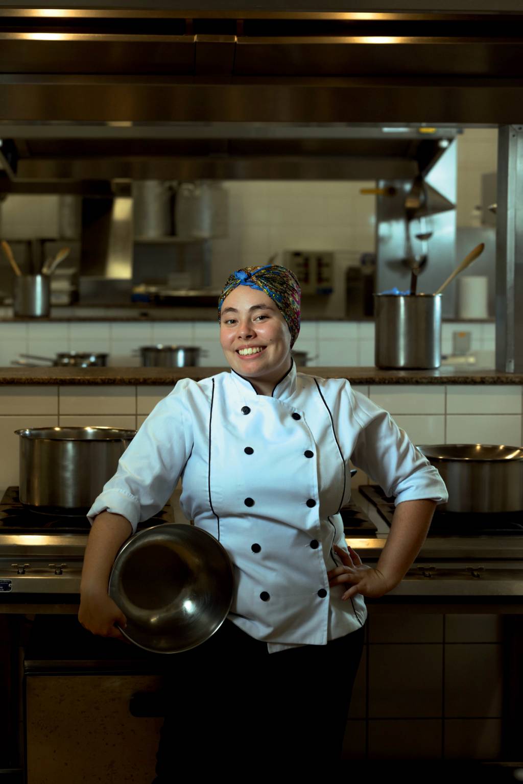 Imagem mostra Bia sorrindo, com roupa de chef de cozinha, segurando um grande recipiente de alumínio na lateral do corpo. Ela está em uma cozinha
