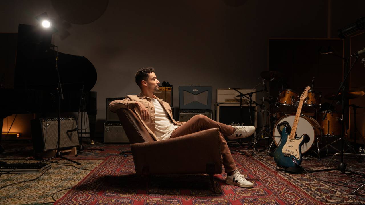 Homem branco posa sentado em estúdio, em poltrona bege, com pernas cruzadas, olhando para o lado e vestindo calça bege e camiseta branca