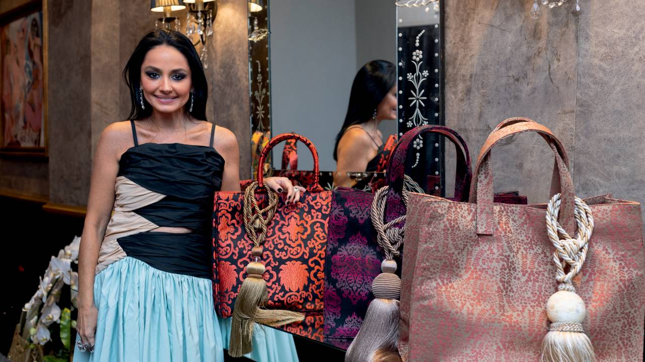 Mulher de cabelos pretos longos e traços árabes posa ao lado de bolsas coloridas. Veste vestido em tons preto, bege e azul bebê.