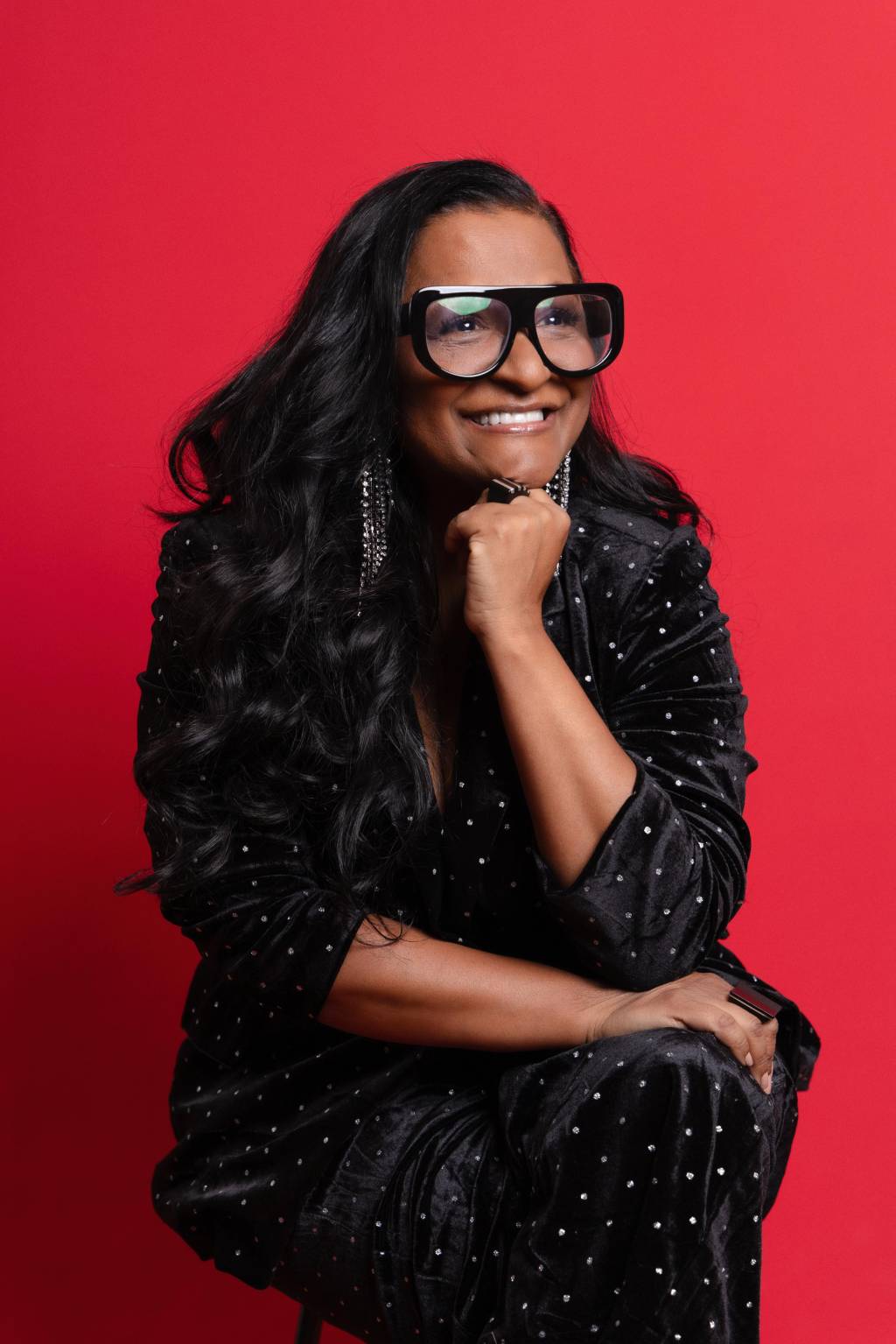 Claudia-Di-Moura-Crédito-Pablo-Grotto Mulher negra com cabelos lisos longos posa em fundo vermelho com roupas pretas, apoiando queixo em um dos pulsos. Sorri e usa óculos de grau pretos