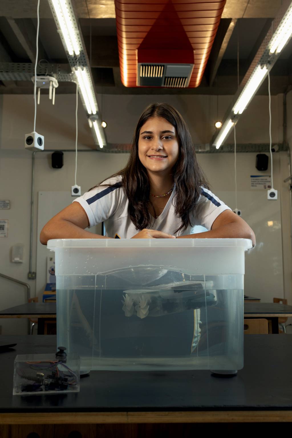 Imagem mostra jovem sentada em cima de mesa com uma caixa de plástico transparente cheia de água e com um dispositivo no interior da caixa