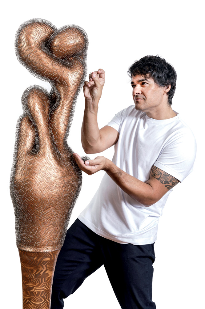 Homem branco de camiseta branca e calça preta segura alfinete e o aponta em direção a escultura em tom de bronze