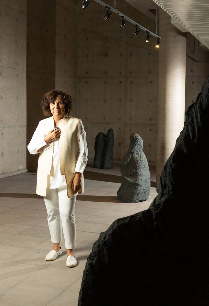 São Paulo, SP – EXPOSIÇÃO DENISE MILAN/USP. Foto: Leo Martins Mulher branca de cabelos castanhos curtos veste roupas brancas e posa de pé envolta por grandes pedras cinza escuro
