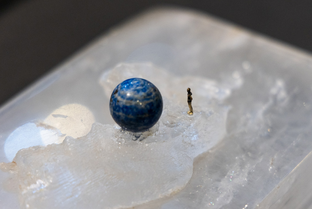 Foto em close exibe bolinha de pedra azul e figura minúscula de ser humano ao lado, ambos em cima de uma pedra lisa branca